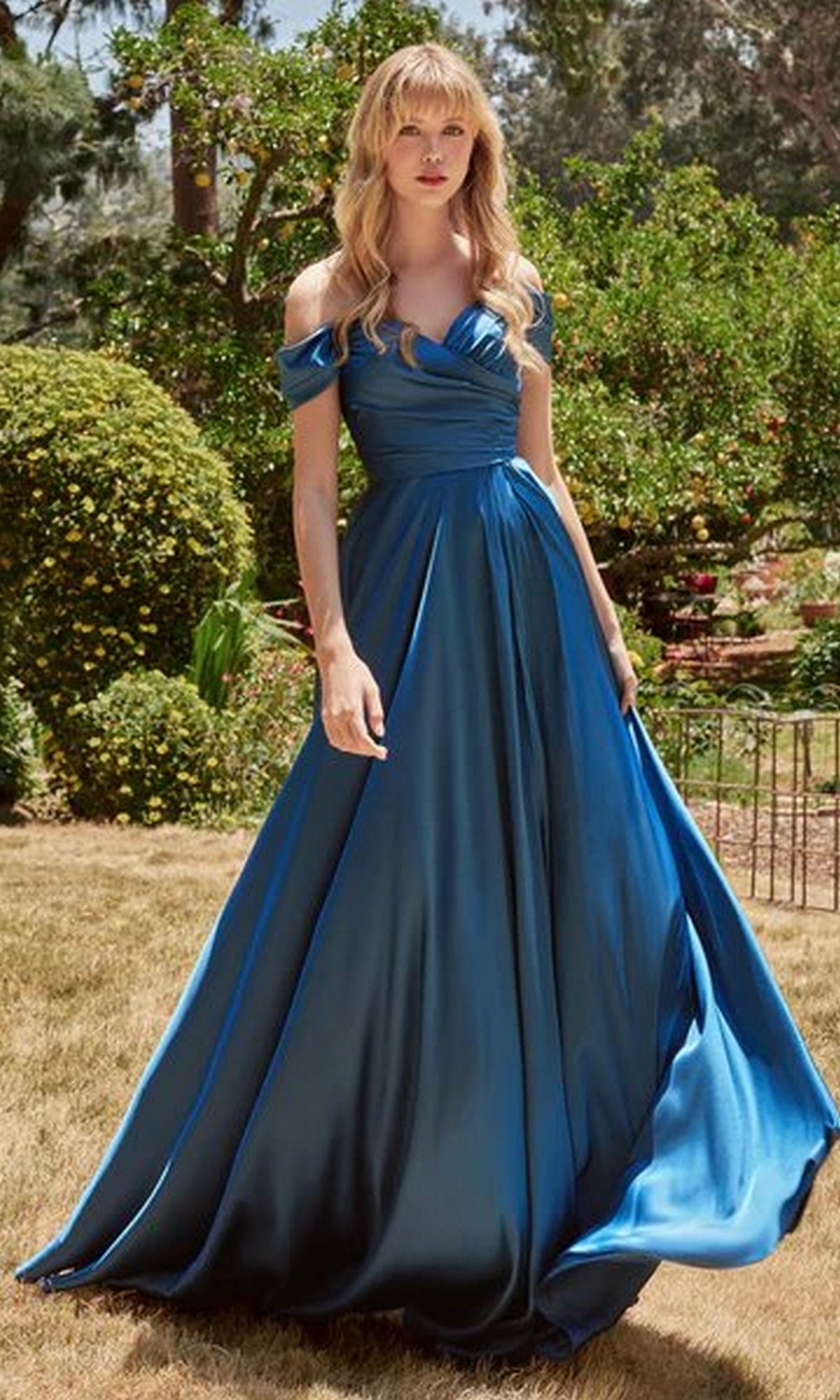 A woman in the Classic Off-Shoulder Long A-Line Prom Dress 7493 stands on dry grass in a garden, surrounded by green bushes and trees.